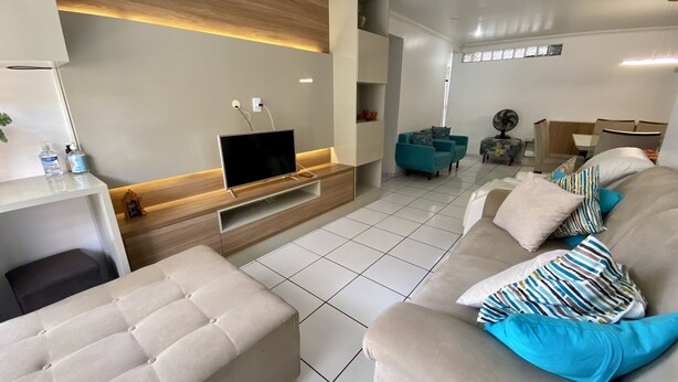 A modern living room with a flat-screen TV on a wooden console, a beige sofa with patterned cushions, and a tiled floor. - 3 quartos a 2 quadras da Praia (Maceió)