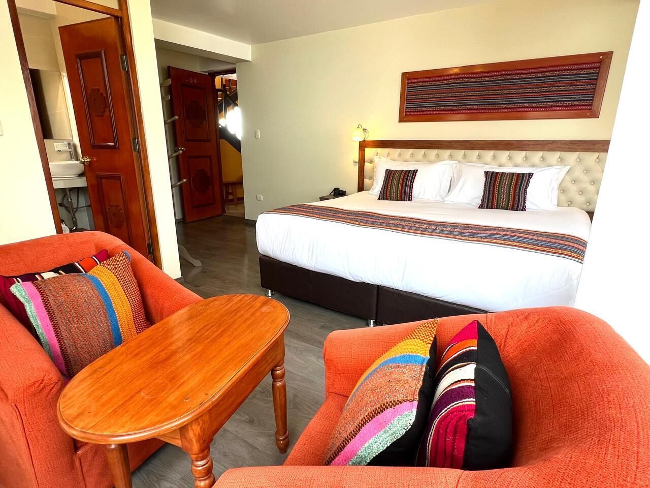 Een hotelkamer met een bed, een houten tafel en twee fauteuils met kleurrijke kussens.