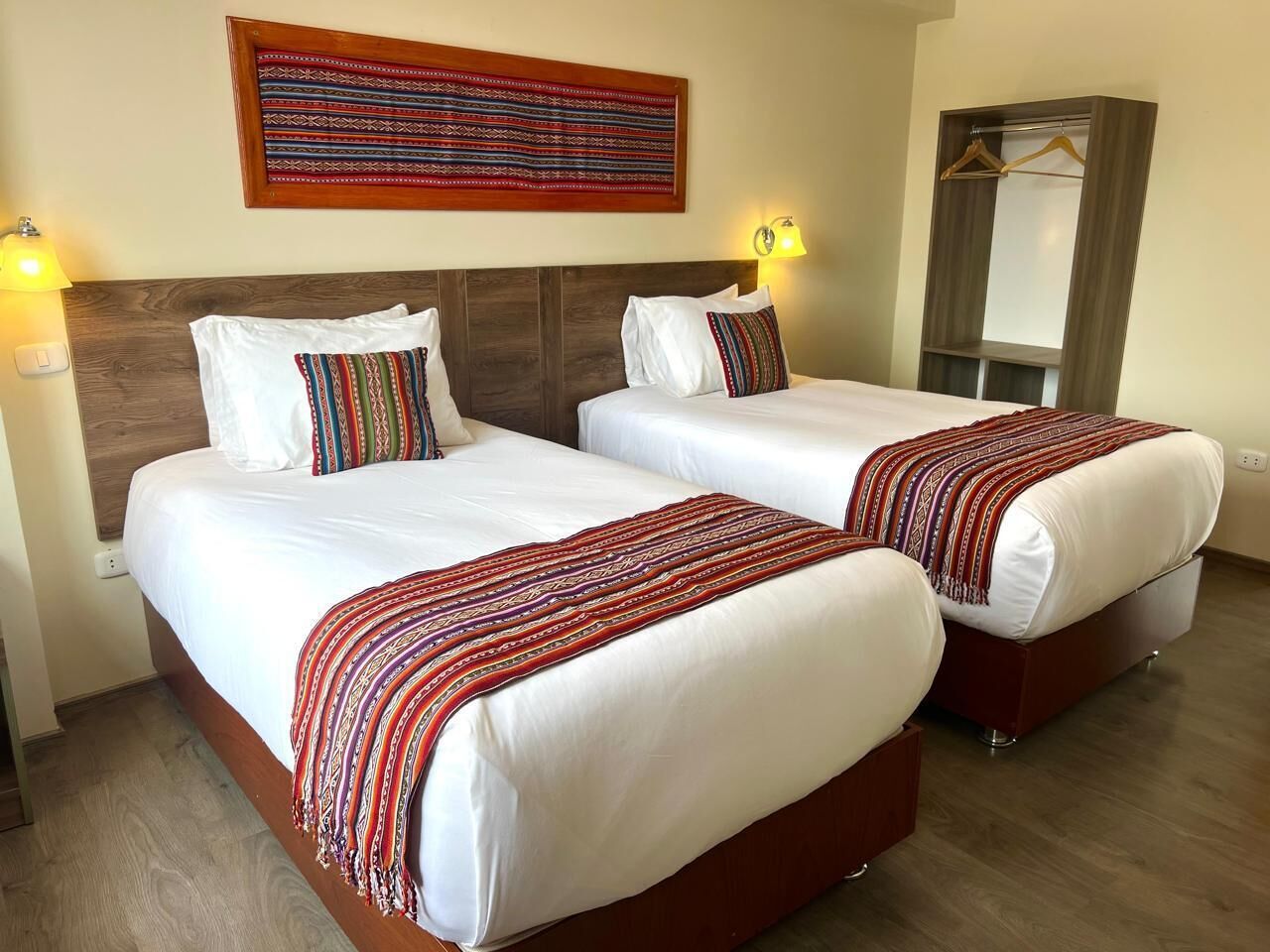 Ein Hotelzimmer mit zwei Betten, jeweils mit weißer Bettwäsche und einer gemusterten Decke. Es gibt ein hölzernes Kopfteil, ein an der Wand befestigtes Kunstwerk und einen Kleiderschrank mit Spiegel.