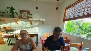A dining area with a red table, plates of food, and a window with a view of greenery. - Pousada Cantinho de taipu (Maraú)