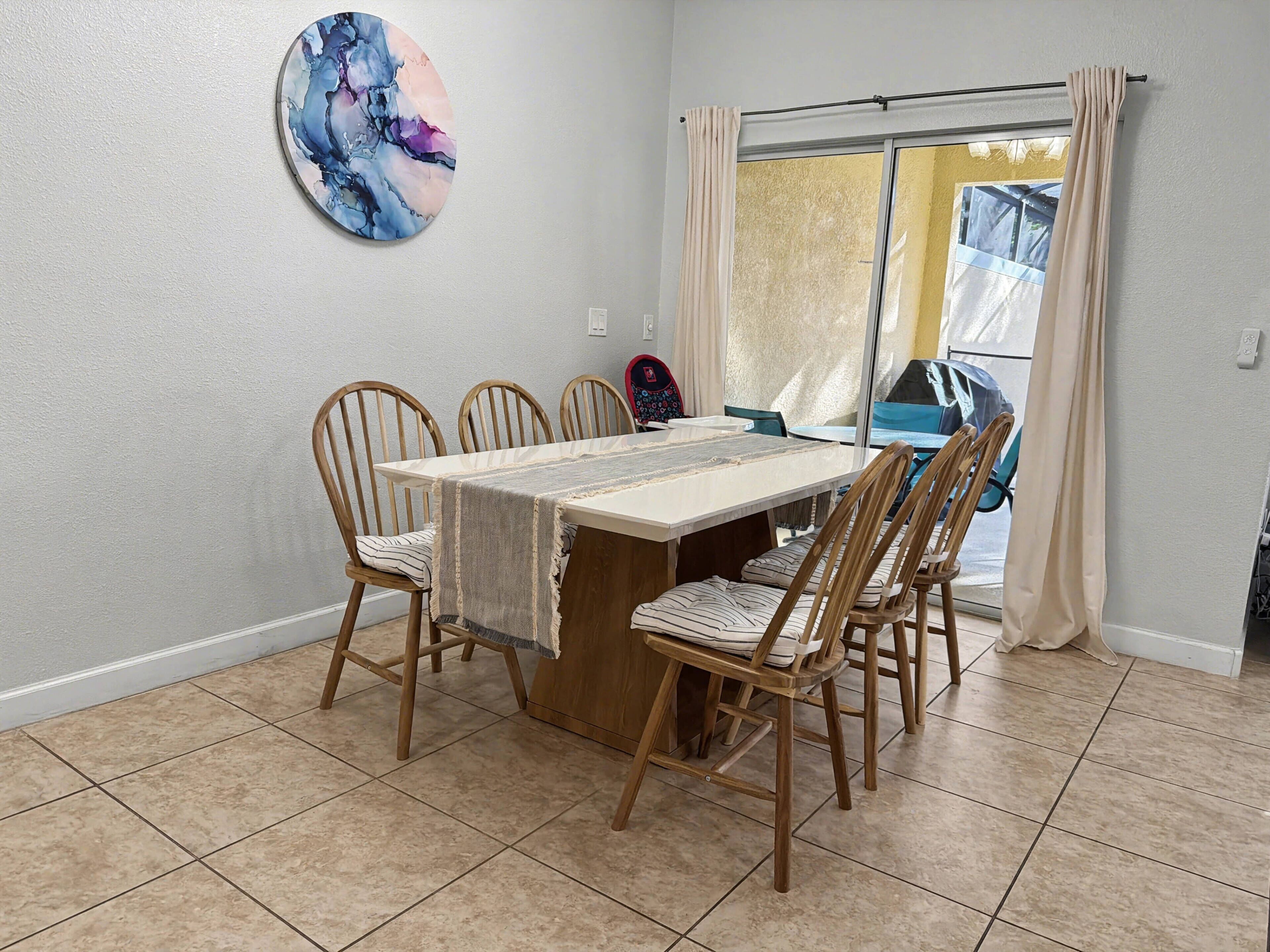 Un espace repas comprenant une table, des chaises, une grande fenêtre avec des rideaux et un tableau accroché au mur.