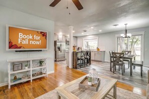 A spacious living room with a large flat-screen TV, wooden flooring, and a dining area with dark chairs and a wooden table.