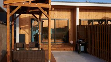 A wooden pergola with wicker furniture, a sliding glass door, and a barbecue grill outside a modern building.