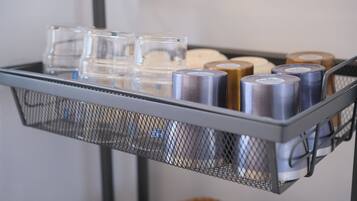 A metal cart with a mesh top holding various items including clear glasses, metal cans, and a brown bag.