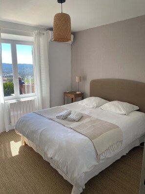 Una cama bien hecha con cabecera, una mesita de noche con lámpara, y vista al exterior a través de una ventana.