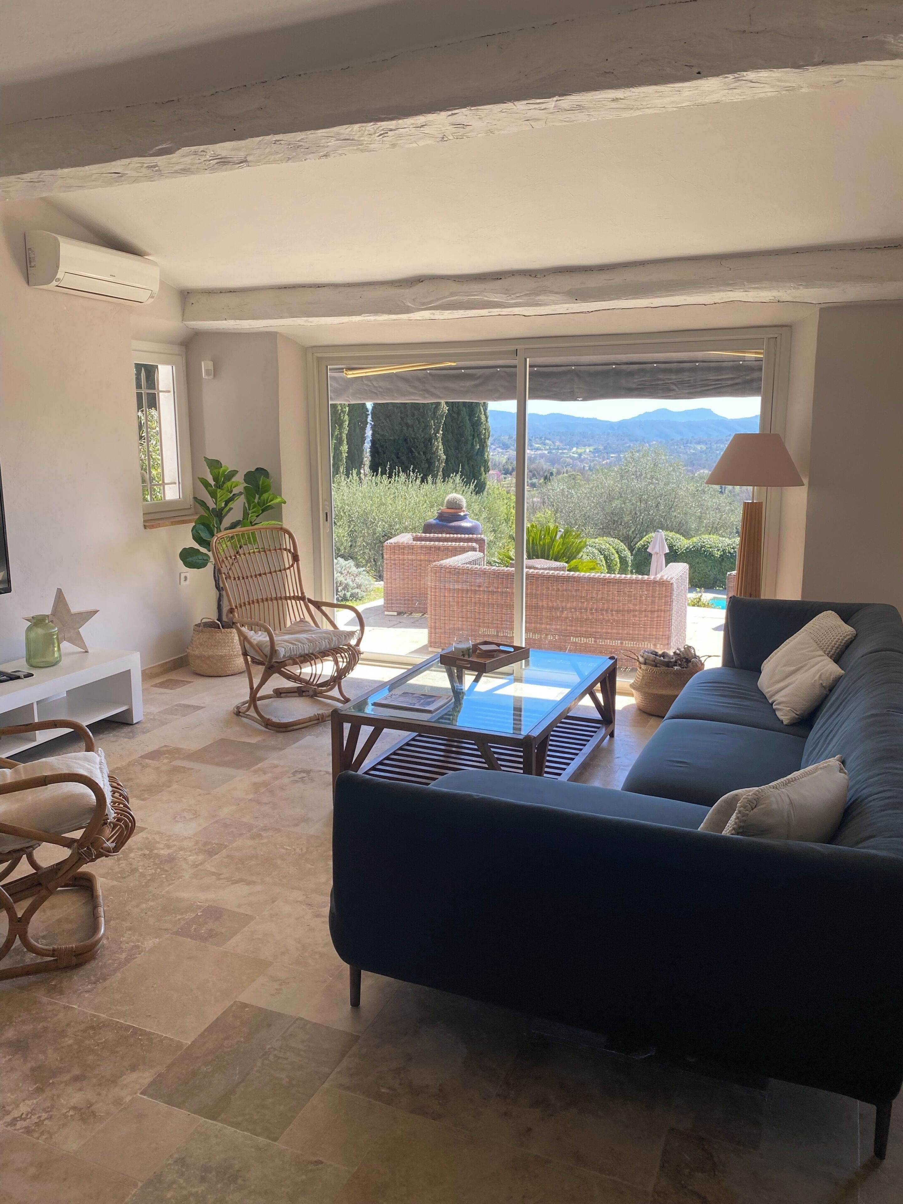 A living room with a dark sofa, wicker chairs, a glass coffee table, and a view of a mountainous landscape through large sliding doors.
