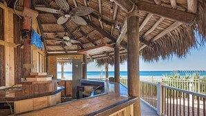 Un bar en la playa con terraza de madera, techo de paja y vista al océano.