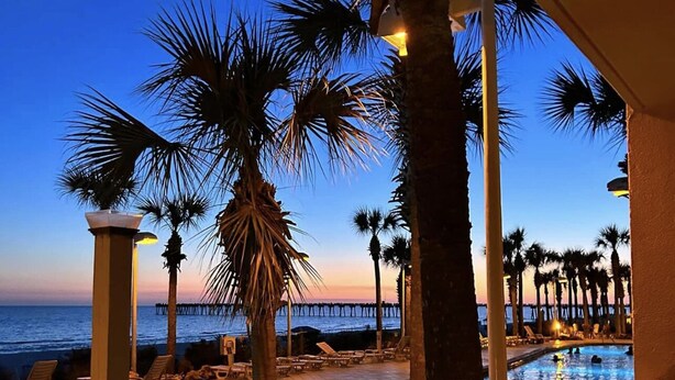 Vista de la piscina al atardecer, con palmeras y un muelle al fondo.
