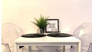 Zona pranzo moderna con tavolo bianco, due sottopiatti neri, un cartello incorniciato, una pianta in vaso e sedie trasparenti.