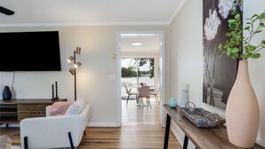 Een moderne woonkamer met een flatscreen televisie, een bank, een eethoek met een tafel en stoelen, en een groot raam met uitzicht op een waterpartij.