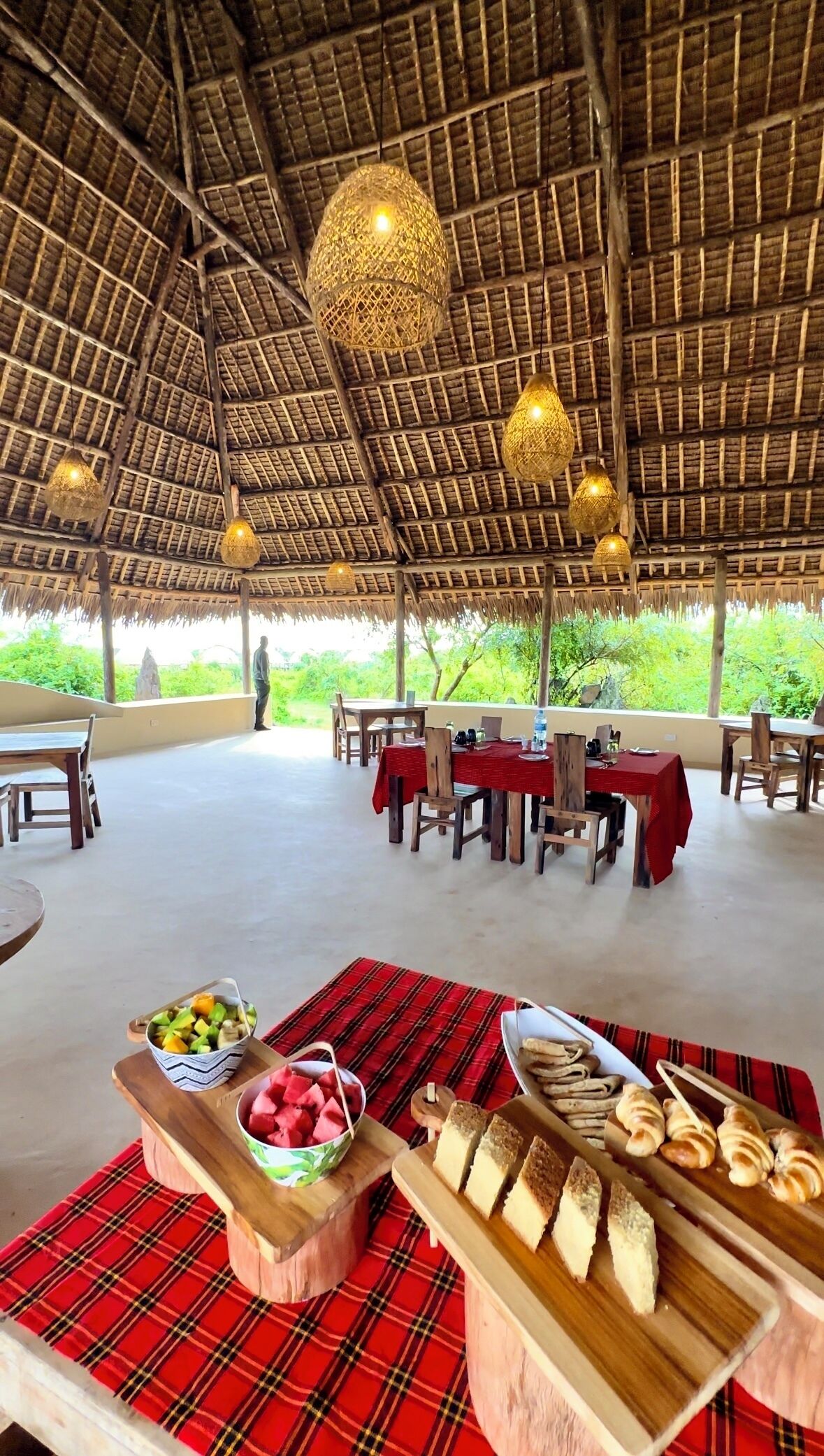 An outdoor dining area with thatched roof, wooden tables, and a buffet with fruits and bread.