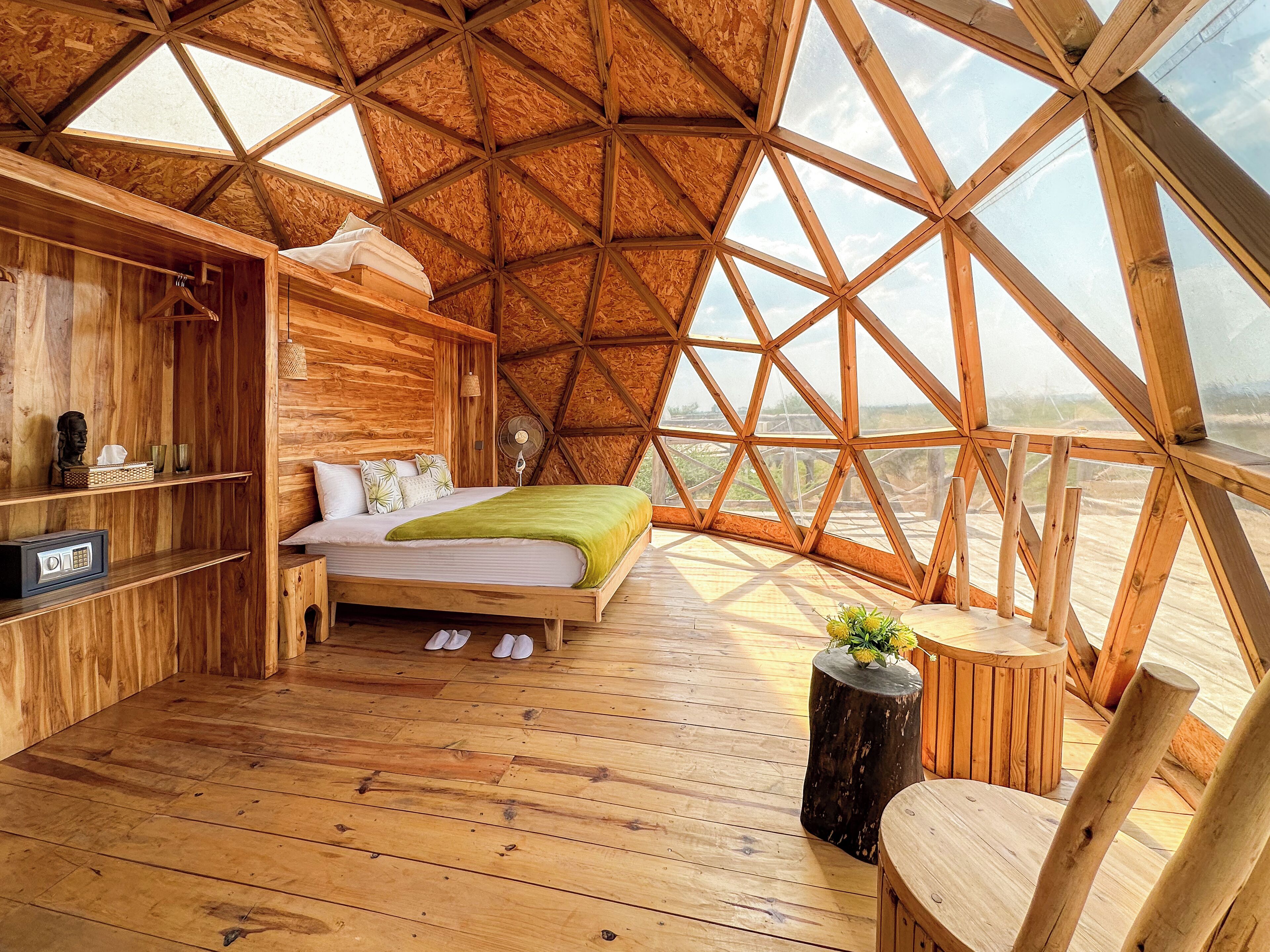 Une chambre confortable avec un plafond en bois et une grande fenêtre, un lit, une table de chevet et une petite table ronde agrémentée d’une plante.