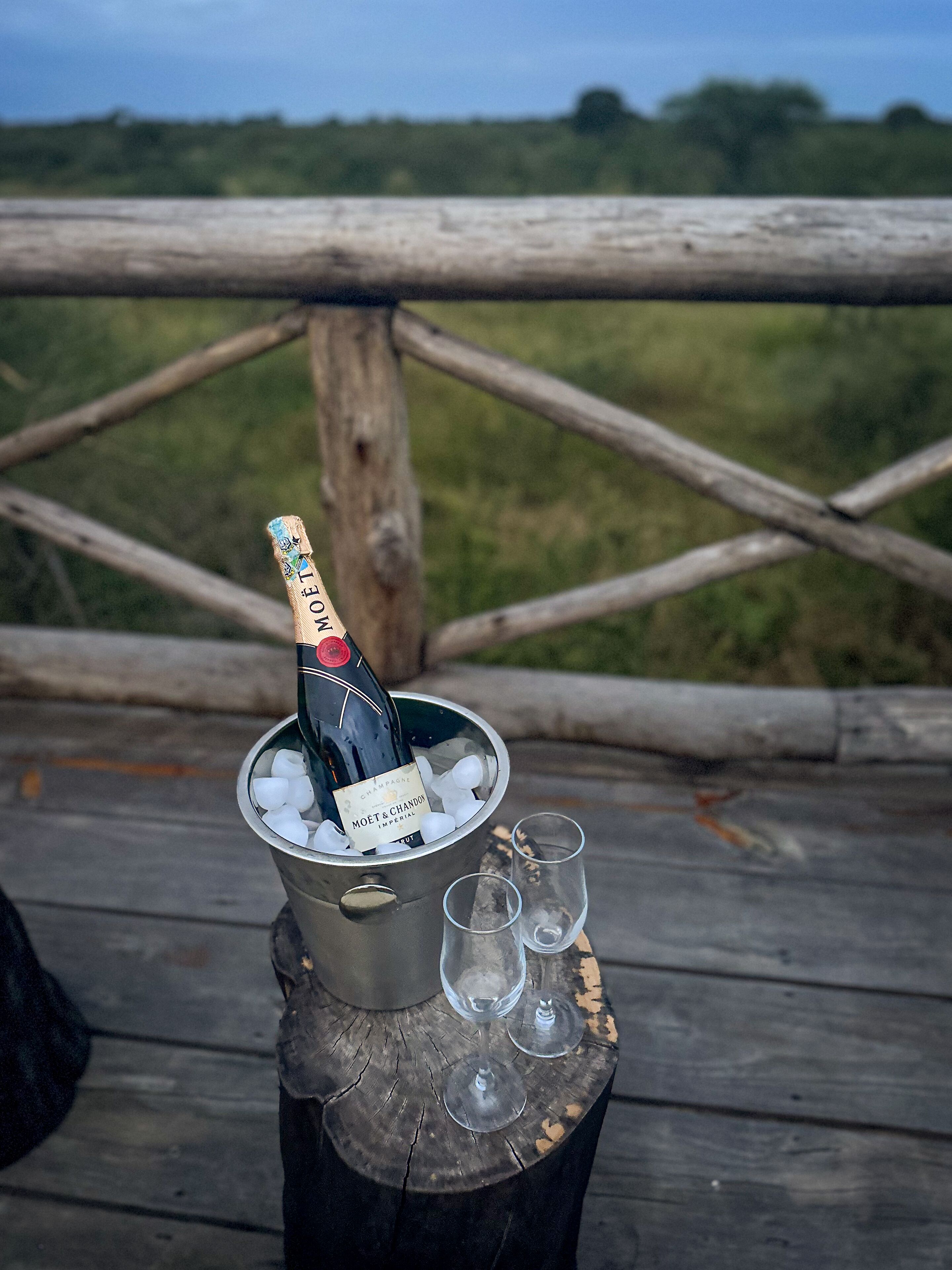 Eine Flasche Moët & Chandon Champagner in einem Metallkübel mit Eis, zwei leere Gläser und ein rustikaler Holztisch auf einer Terrasse mit einer Holzgeländer im Hintergrund.
