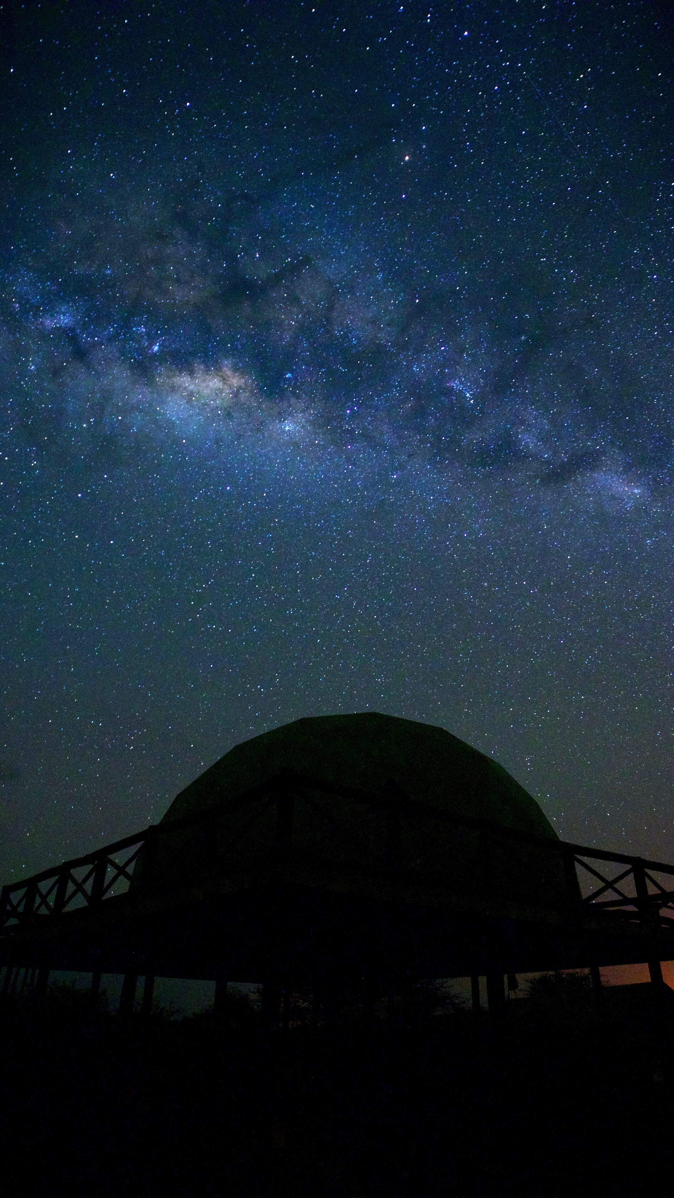 Cielo nocturno lleno de estrellas y la Vía Láctea sobre una estructura oscura con techo.