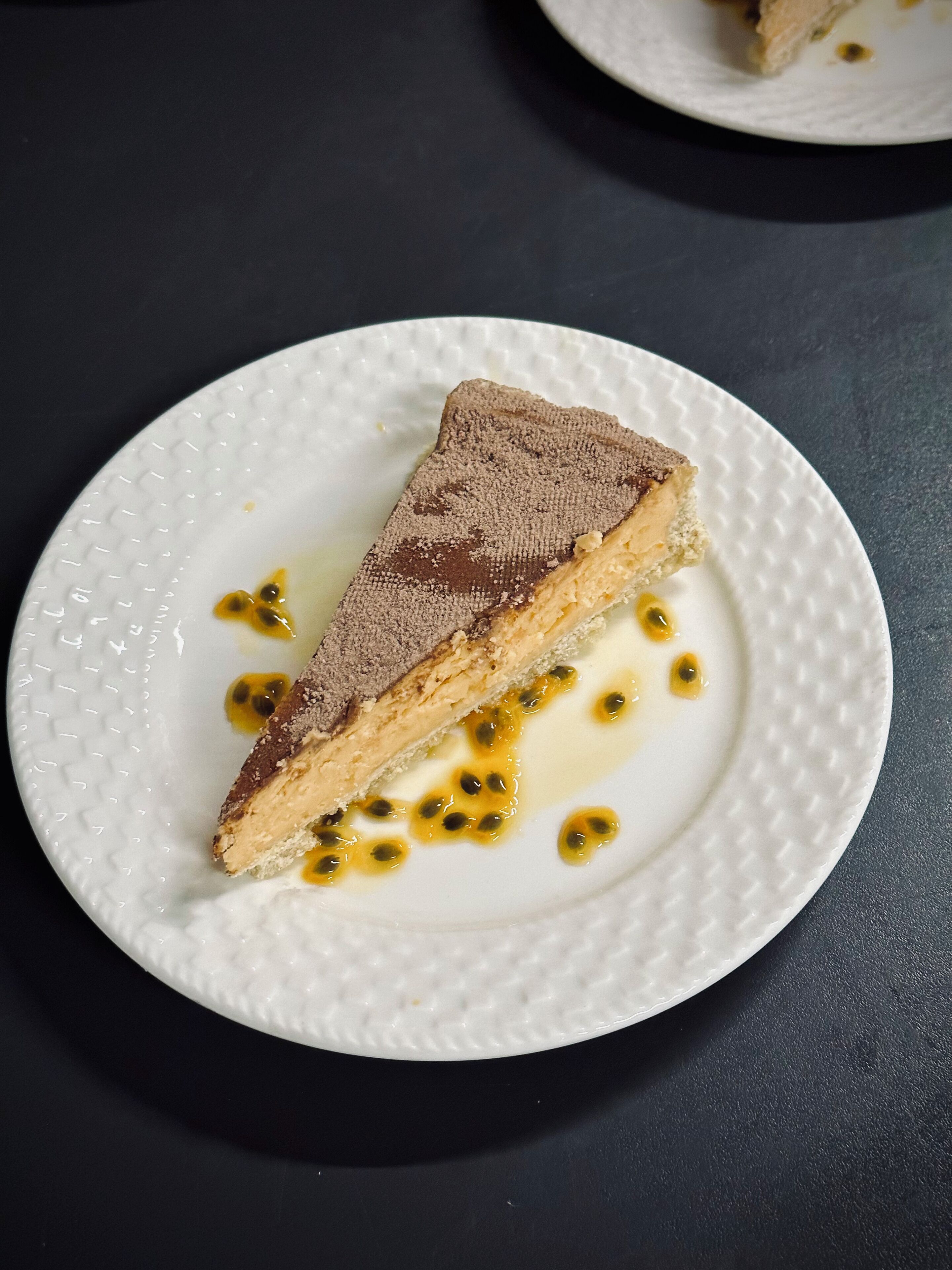 Une part d’un dessert à plusieurs couches, présentée sur une assiette blanche, le tout sur un fond sombre.