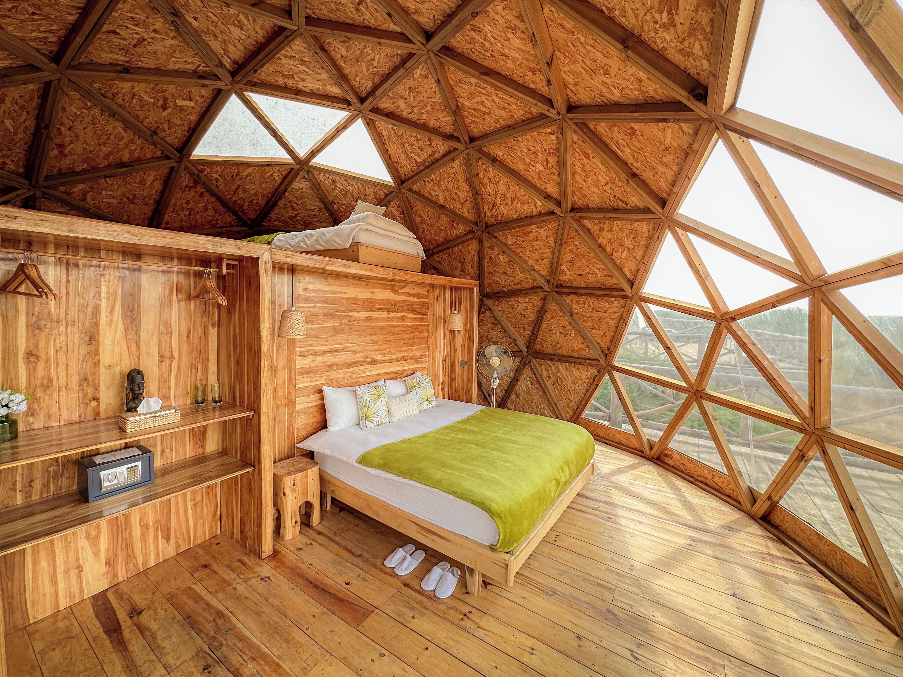 Een houten kamer met een hoge plafond, een bed met een groene deken, een houten dressoir en een klein krukje.
