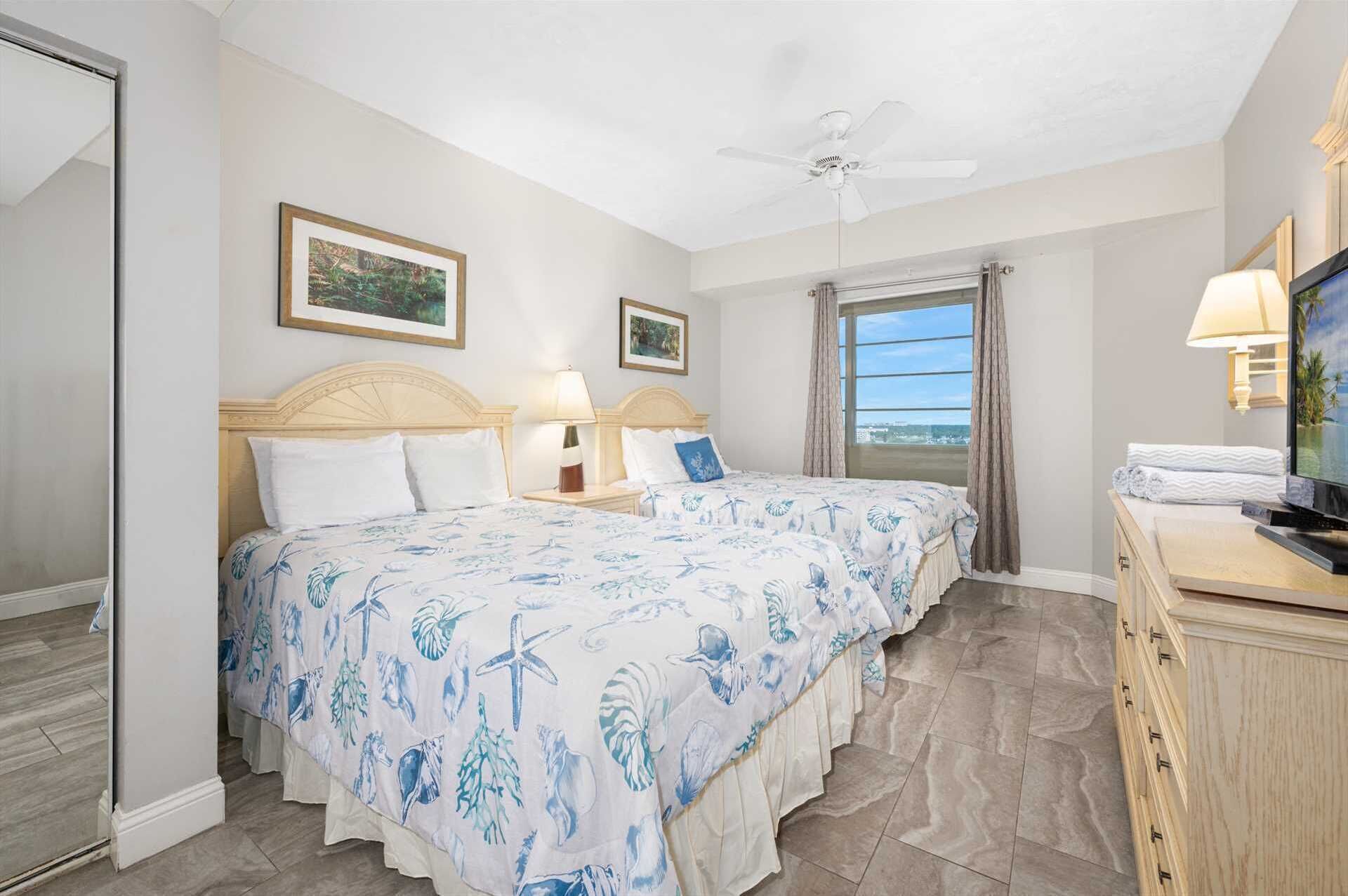 A hotel room with two beds, a ceiling fan, a TV, and a view of the ocean.