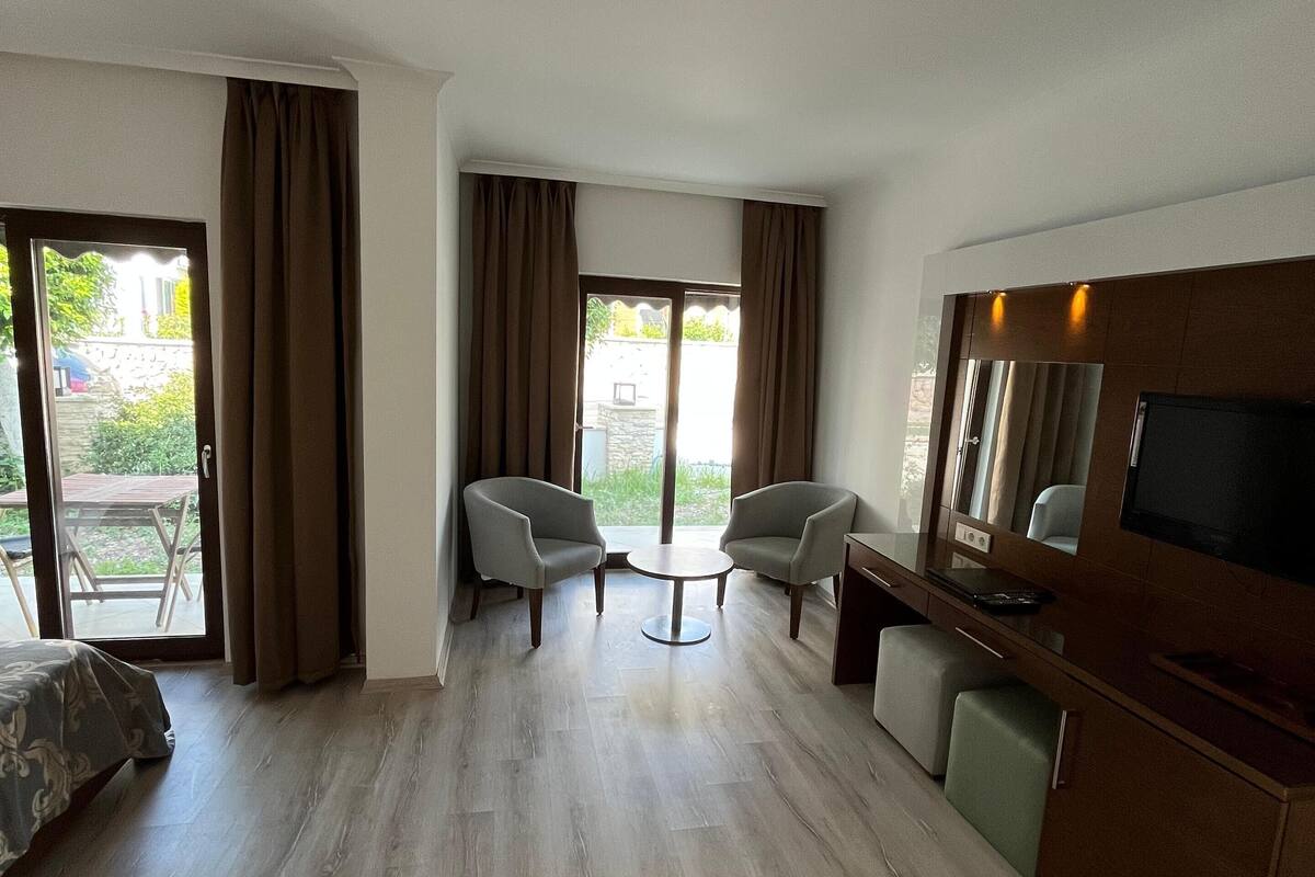 A hotel room with a bed, a desk with a TV, two chairs, a small table, and a sliding glass door to the outside.