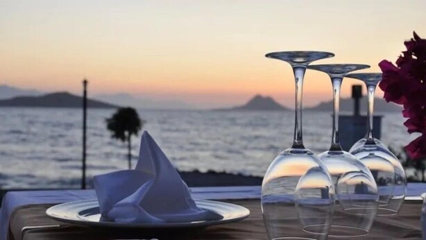 Une table dressée avec des verres à vin et une serviette pliée, offrant une vue sur la côte et un coucher de soleil.