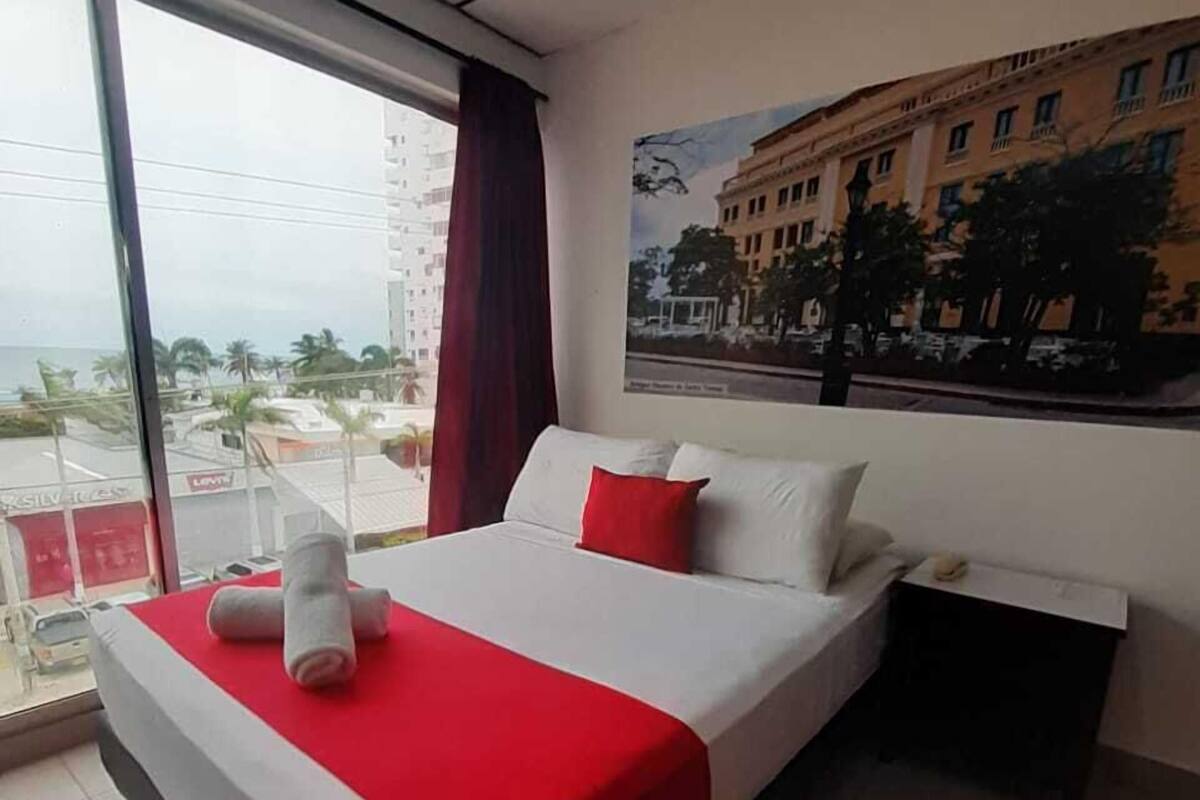 A hotel room with a large bed, a nightstand, a window with a view of buildings and palm trees, and a poster on the wall.