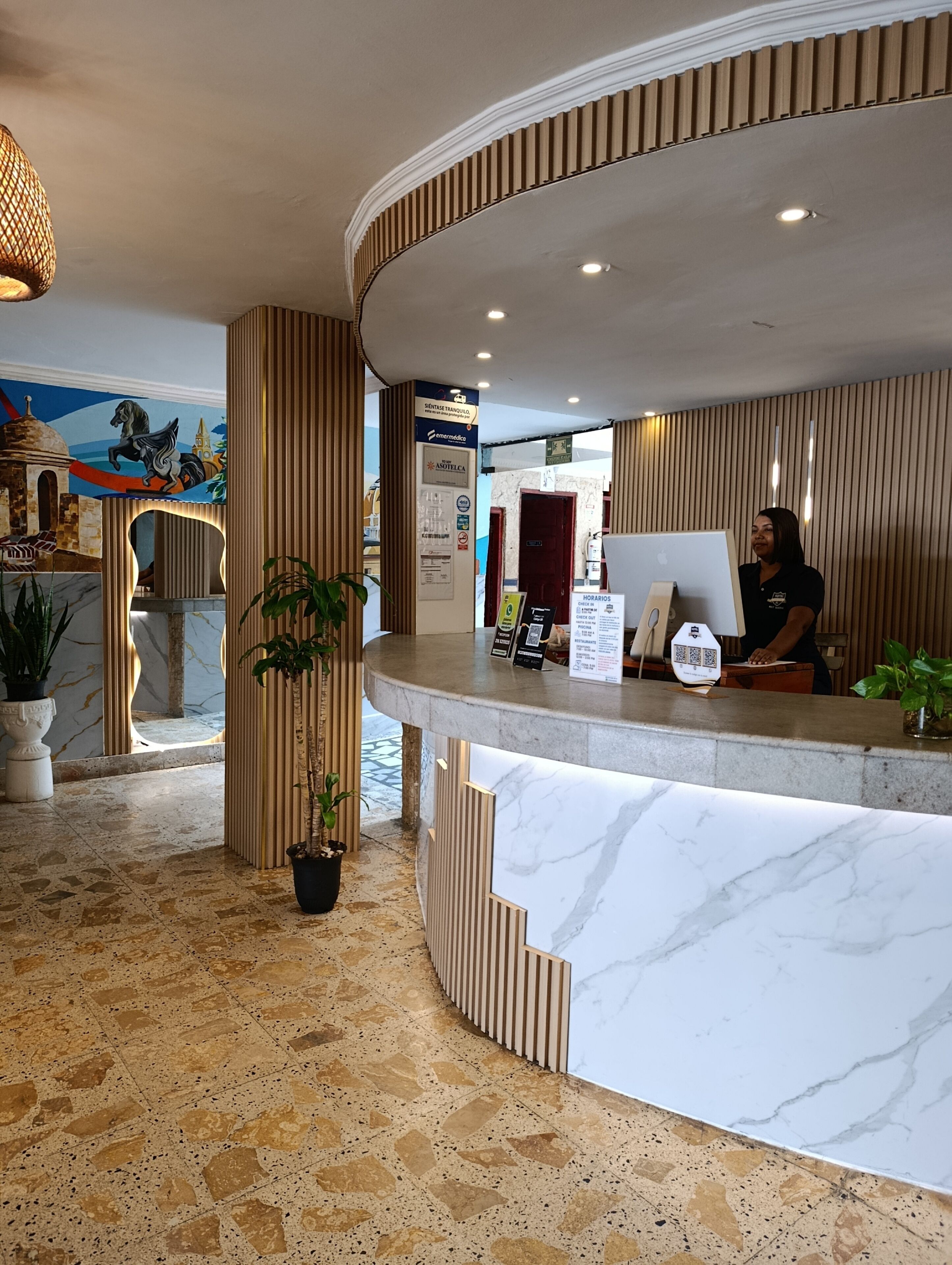 A hotel reception area with a curved marble counter, a receptionist, and a wall with a mural.