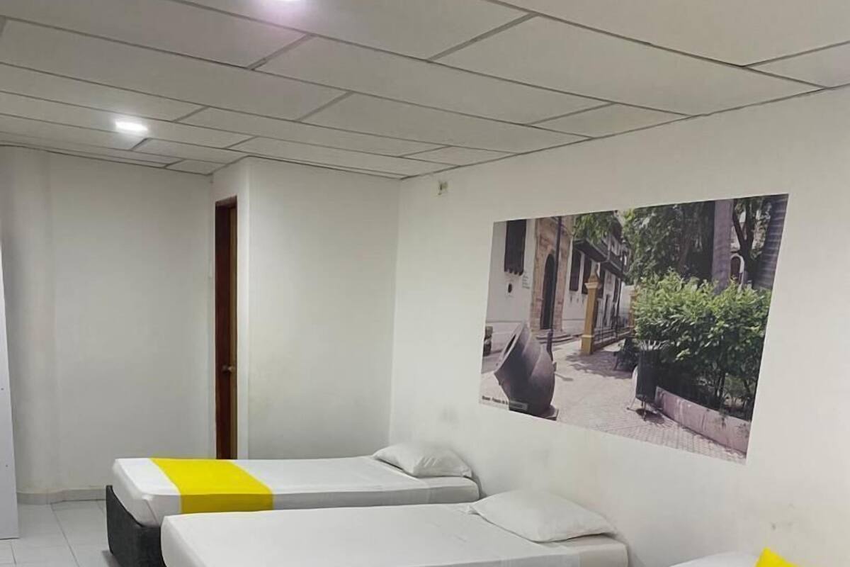 A hotel room with three beds, each with a white mattress and a yellow accent. There is a wooden table and a plastic chair in the room.