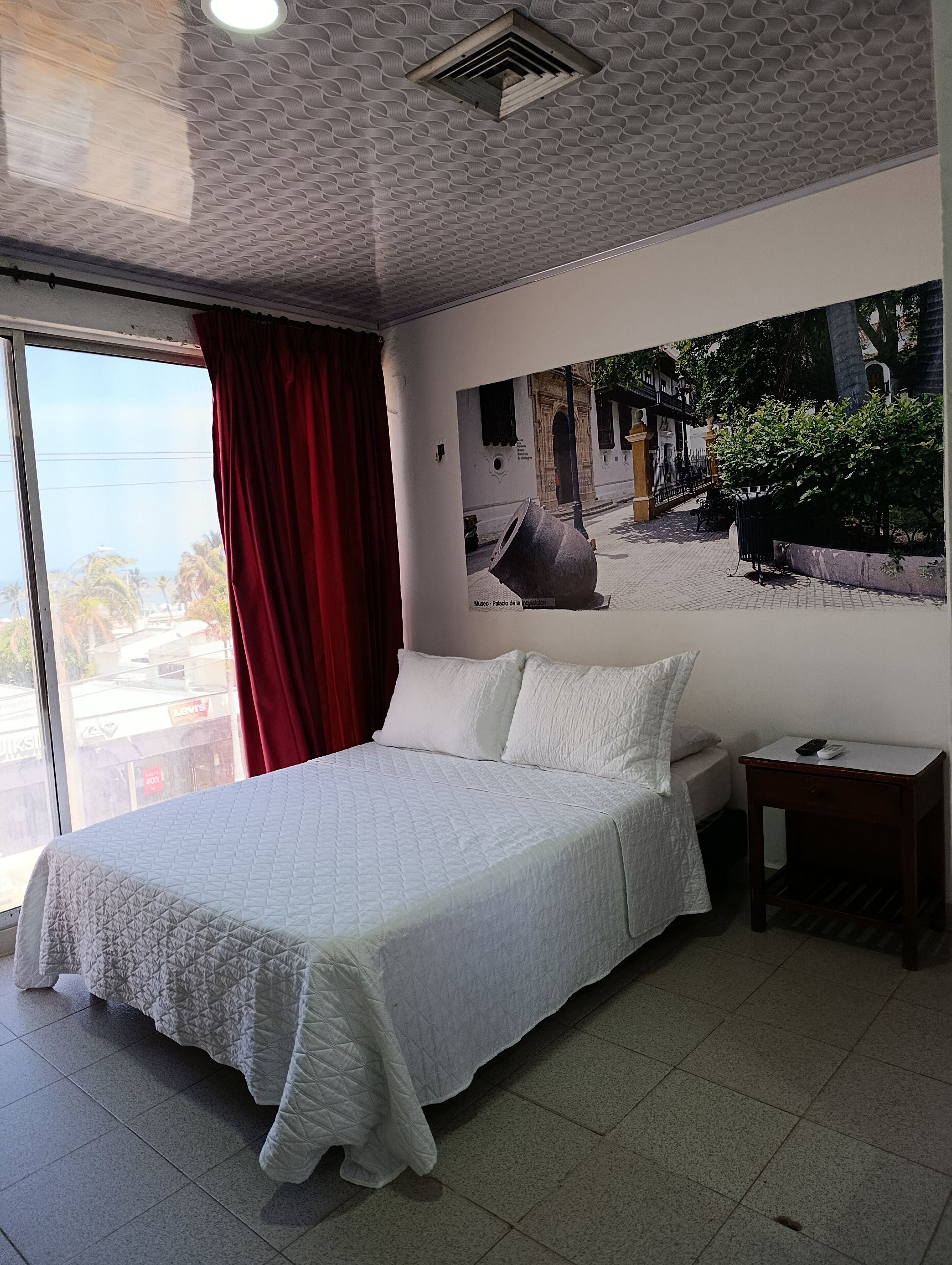 Une chambre à coucher avec un grand lit, une table de chevet, une fenêtre avec des rideaux rouges et un tableau accroché au mur.