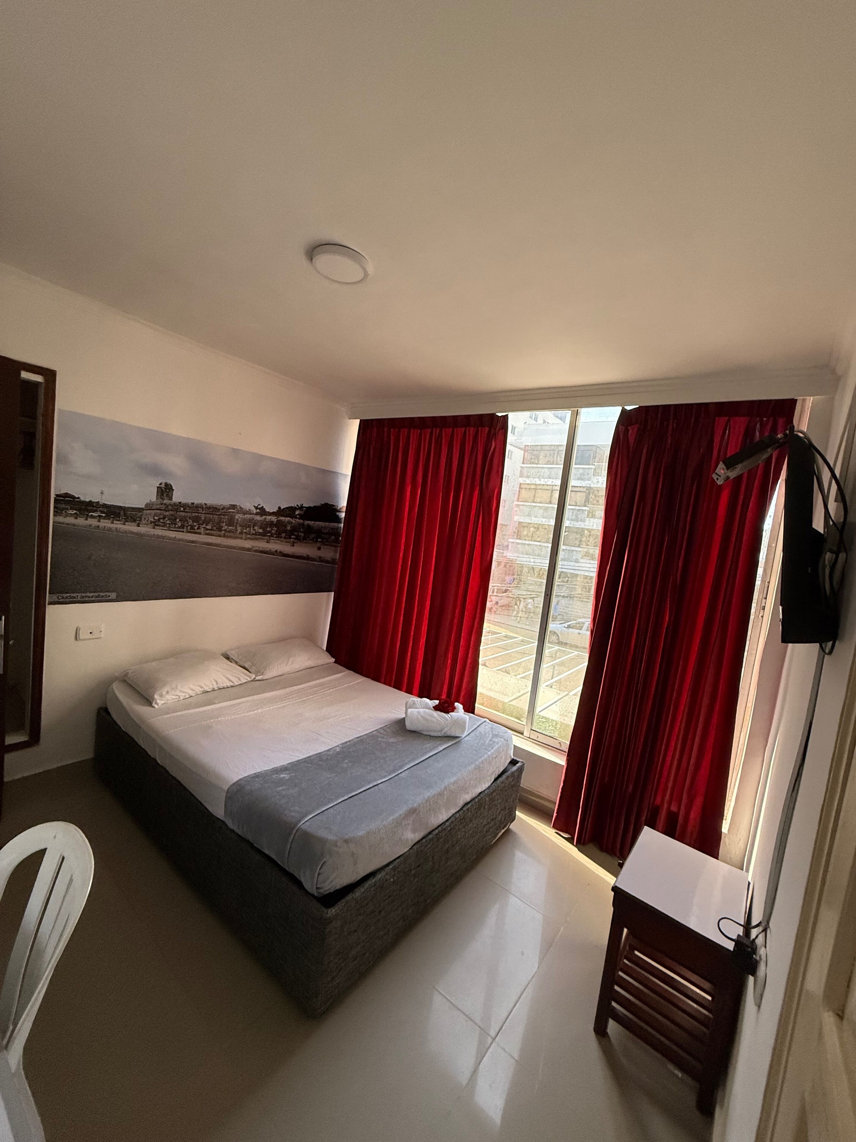 Habitación de hotel con cama, escritorio, silla, televisor y un amplio ventanal con cortinas rojas.