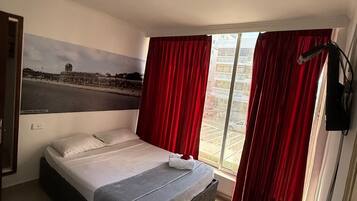 Habitación de hotel con cama, escritorio, silla, televisor y un amplio ventanal con cortinas rojas.