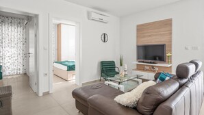 Uma sala de estar moderna com um sofá de couro marrom, uma mesa de centro de vidro, uma poltrona verde, uma TV em uma parede de madeira e uma cama ao fundo.