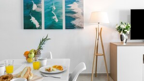 Un coin repas moderne avec une table blanche dressée pour le petit-déjeuner, comprenant des fruits, du pain et du jus. Au mur, on peut admirer trois tableaux encadrés représentant des vagues océaniques. Une lampe sur pied et un téléviseur posé sur un meuble complètent l’ensemble.