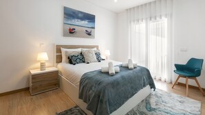 Une chambre à coucher moderne avec un lit, des tables de chevet, une chaise et un tableau représentant des bateaux sur une plage.