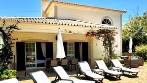 A building with a tiled roof, a covered patio with lounge chairs and umbrellas, and a garden with flowering plants. - Attractive Family Villa, A/C, Private Pool - 5mins drive to the Iconic Beach at Praia da Marinha! (Lagoa)