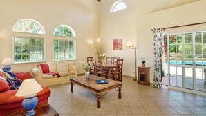 A spacious living room with a high ceiling, large windows, a dining area, and a view of a swimming pool. - Attractive Family Villa, A/C, Private Pool - 5mins drive to the Iconic Beach at Praia da Marinha! (Lagoa)