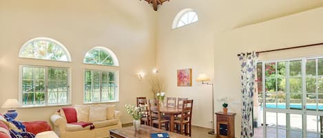 A spacious living room with a high ceiling, large windows, a dining area, and a view of a swimming pool.