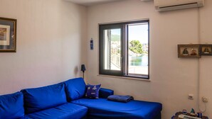 A room with a blue sofa, a window with a view of buildings and water, and an air conditioning unit on the wall. - 3 bedroom beautiful apartment in Klek (Klek)