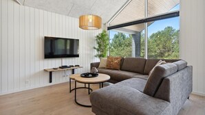 Ein modernes Wohnzimmer mit einer Eckcouch, einem runden Couchtisch, einem an der Wand befestigten Flachbildfernseher und einem großen Fenster mit Blick auf Bäume.
