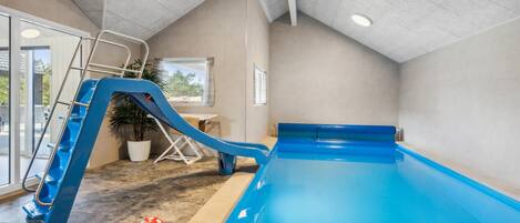 An indoor swimming pool with a blue slide, a ladder, and a red ball on the floor.