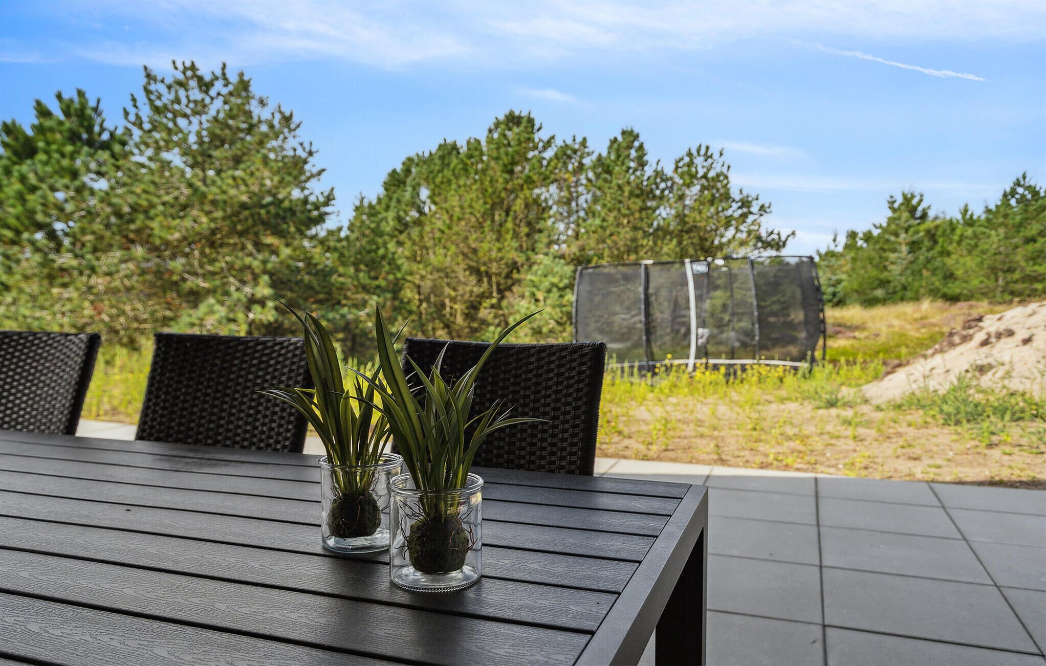 A wooden table with two glass vases containing plants, set outdoors with a view of trees and a fence in the background.
