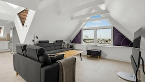 Un loft moderno con techo inclinado, un sofá gris, una mesa de centro de madera y vistas a un puerto deportivo a través de amplios ventanales.