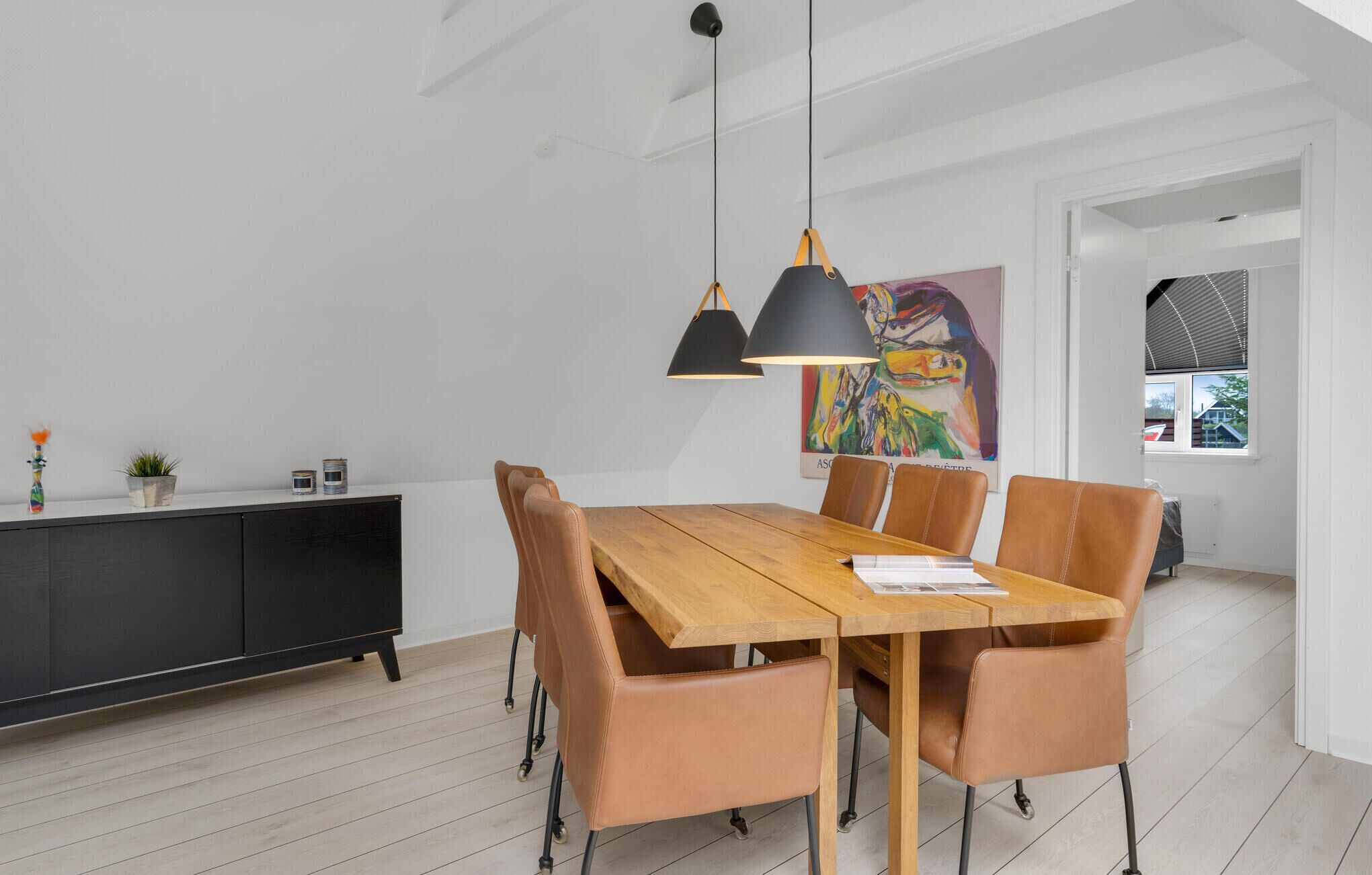 Un comedor moderno con mesa de madera, sillas de cuero, lámparas colgantes negras y un armario negro decorado con objetos.