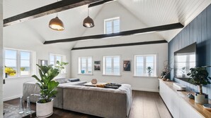 Une pièce spacieuse avec un grand lit, de nombreuses fenêtres offrant une vue sur l’océan, des poutres en bois au plafond et un coin cuisine doté d’un plan de travail en bois.