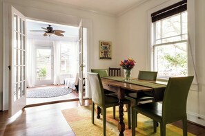 A dining area with a wooden table, green chairs, and a vase of flowers. There is a ceiling fan, a window with blinds, and a door leading to an outdoor area.