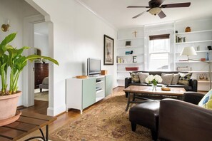 A living room with a sofa, coffee table, and a television on a stand. There is a bookshelf with books and decorative items. A potted plant is visible in the corner.