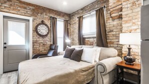 Un dormitorio con pared de ladrillo, una cama con cubierta color beige, un reloj, una silla y una mesita de noche con una lámpara.