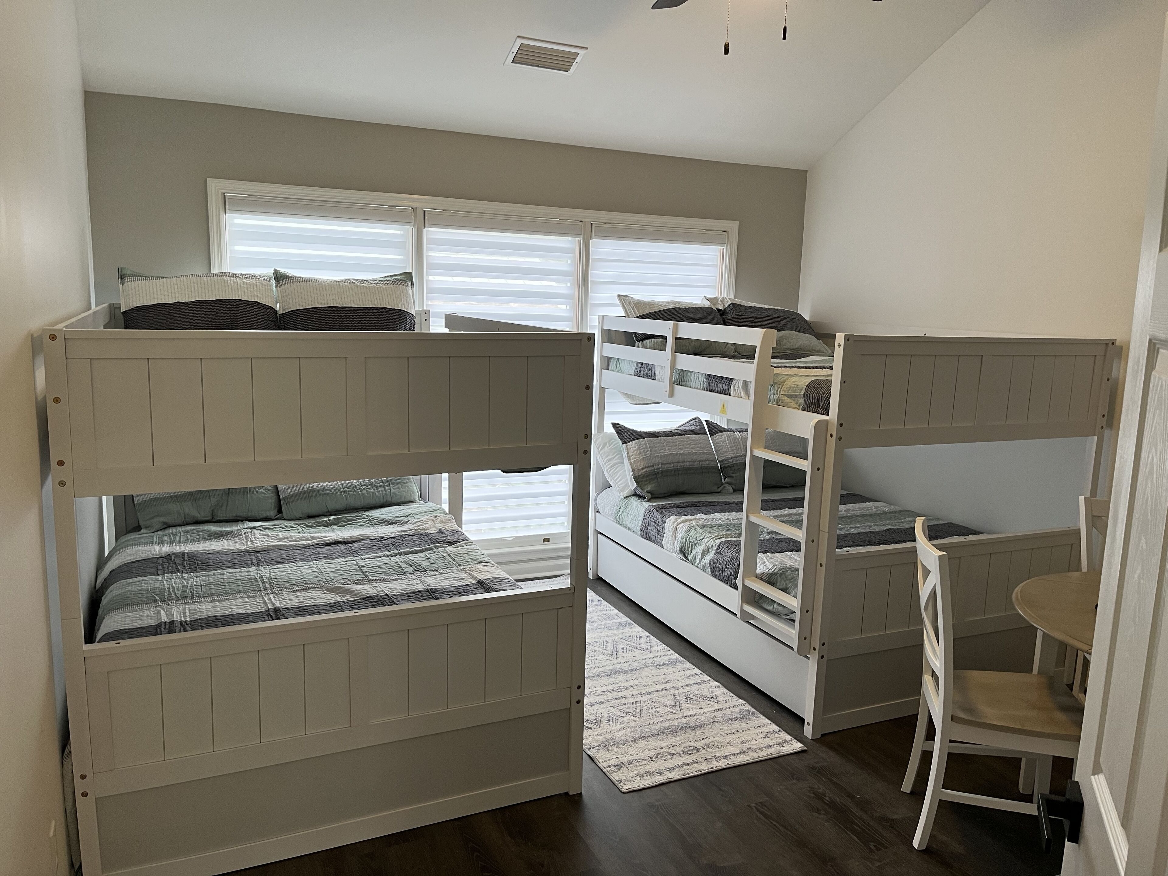 Een slaapkamer met stapelbed, bureau en stoel, een raam met jaloezieën, en een vloerkleed.
