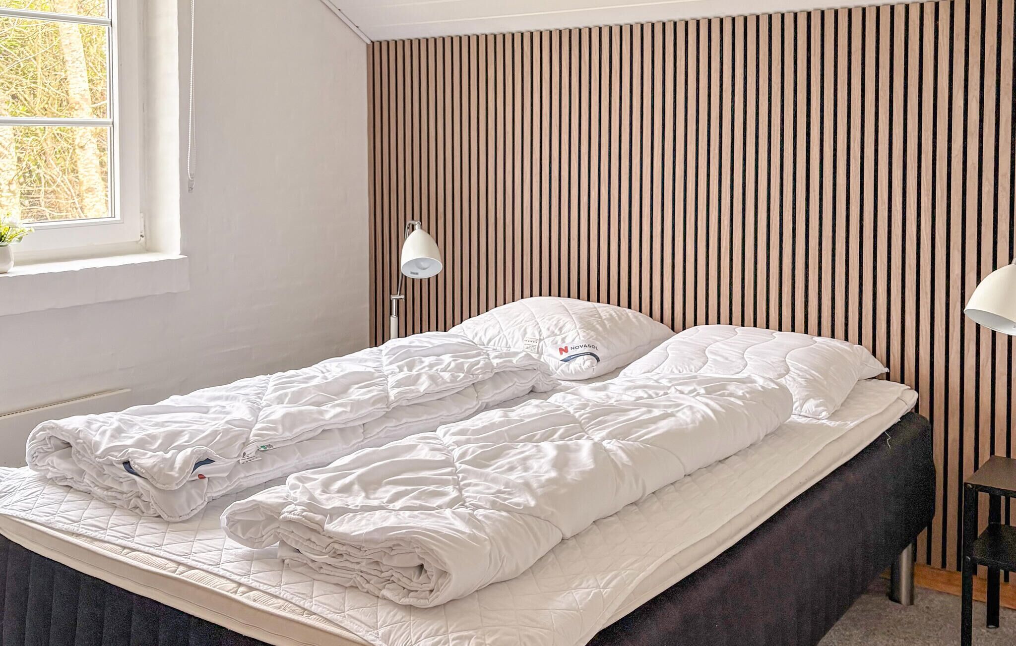 A neatly made bed with a white duvet and pillows, a bedside lamp, and a window with a view of trees.