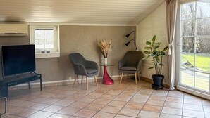 A living room with a television, two chairs, a red vase with dried plants, a potted plant, and a large window with curtains. - Gorgeous home in Hemmet with WiFi (Hemmet)