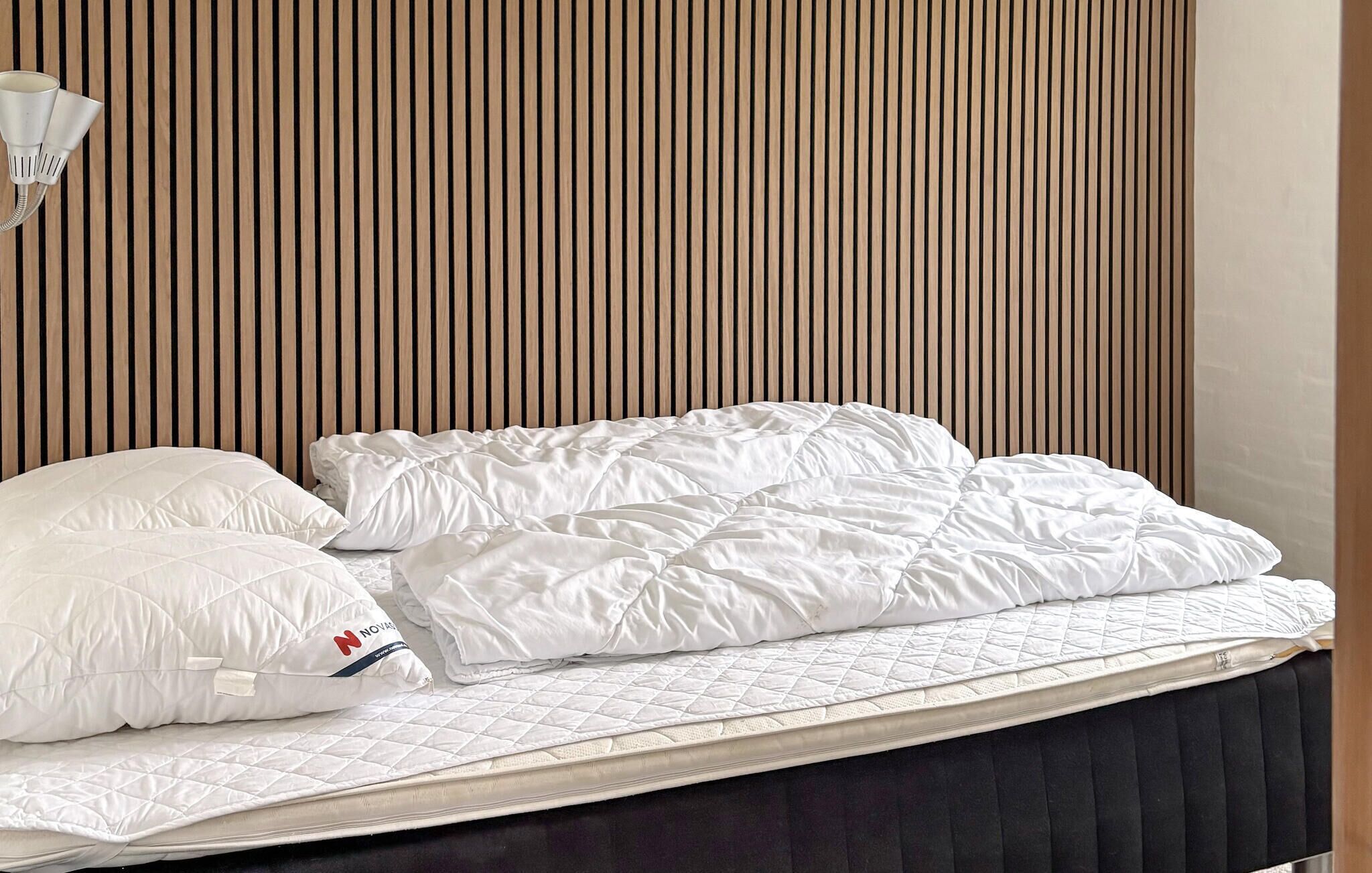 Een net opgemaakt bed met witte lakens en een kussen met een logo, voor een muur met verticale houten latten.