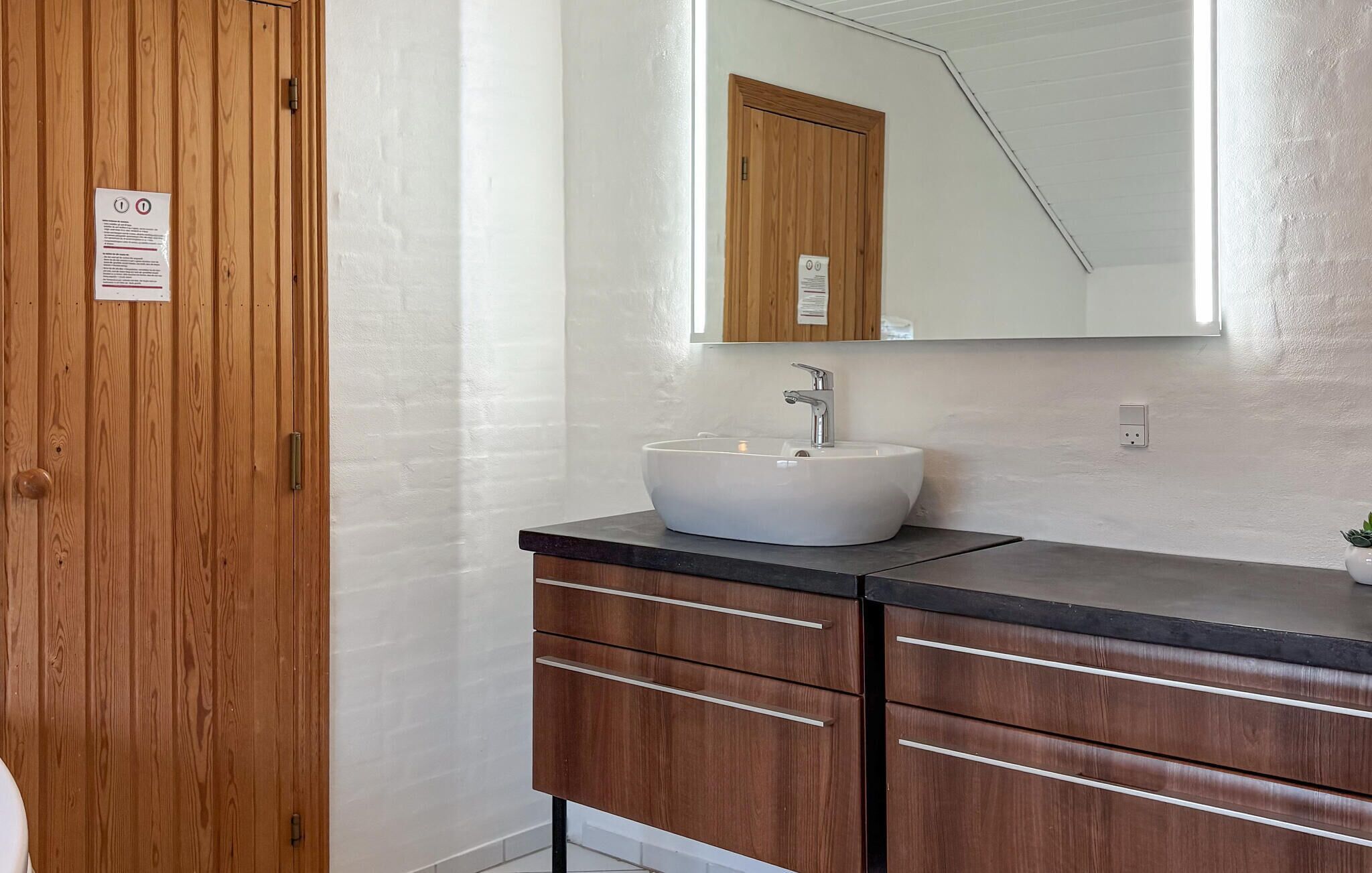 A bathroom with a wooden door, a white sink with a modern faucet, a mirror above the sink, and a dark countertop.
